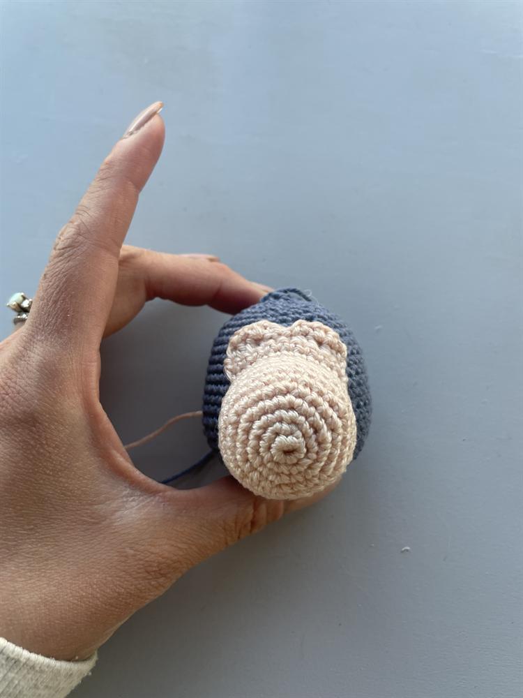 A hand holding a small crocheted slate-blue amigurumi head, showing the cream spiral snout stitched to its front