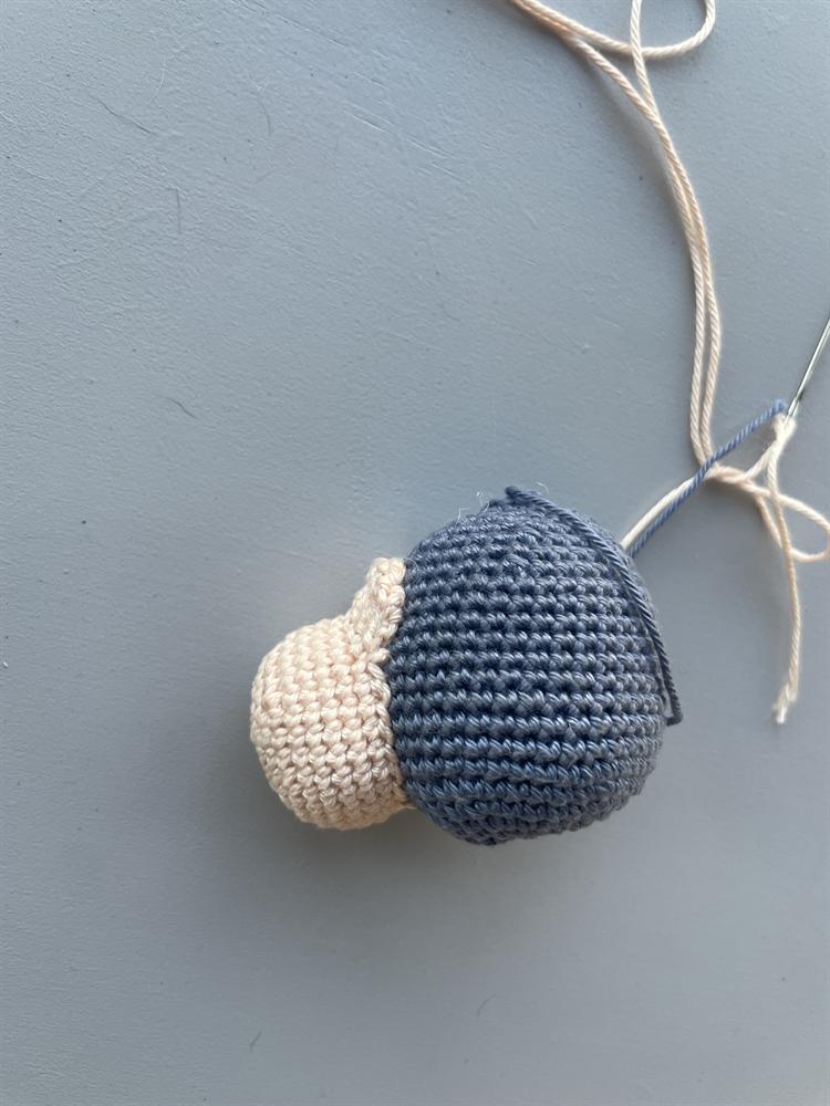 A small crocheted amigurumi head in slate-blue yarn with a cream snout, lying on a gray surface beside a tapestry needle threaded with cream yarn