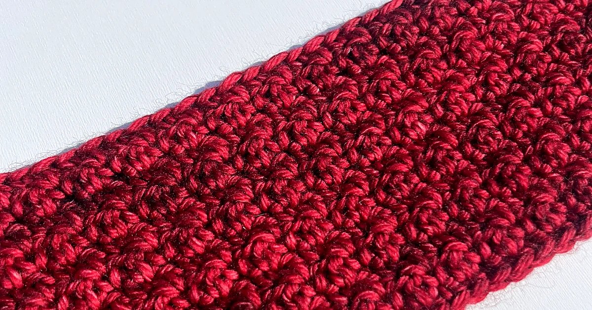 A close up of a strip of dark red crochet fabric worked in griddle stitch, lying on a white surface, with the nubbly alternating texture running in diagonal rows